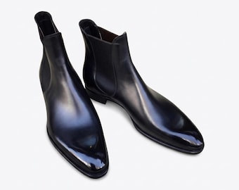 May include: A pair of black leather Chelsea boots with a glossy finish. The boots have a sleek, pointed toe design and elastic side panels. The footwear is displayed on a white background, highlighting the polished appearance of the leather.