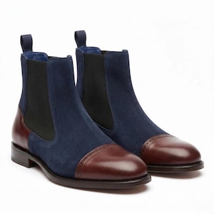 Könnte beinhalten: Ein Paar blaue Chelsea-Stiefel aus Wildleder mit braunen Lederkappen und Fersenakzenten. Die Stiefel haben elastische Seiteneinsätze und Zuglaschen. Das Schuhwerk wird auf weißem Hintergrund präsentiert.
