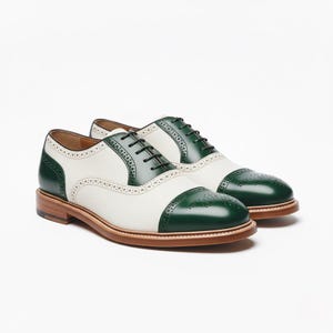 Könnte beinhalten: Zweifarbige Oxford-Schuhe in Weiß und Grün. Die Schuhe haben grüne Zehenkappen, Fersenteile und Ösen, mit weißen Lederoberteilen. Sie haben braune Sohlen und schwarze Schnürsenkel. Auf weißem Hintergrund präsentiert.