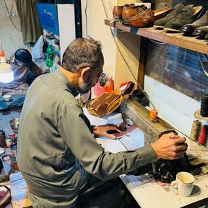 May include: A person in a grey long-sleeved shirt operates a vintage industrial sewing machine to stitch leather pieces. The workspace features various spools of colored thread, finished leather shoes on a shelf, and scattered footwear.