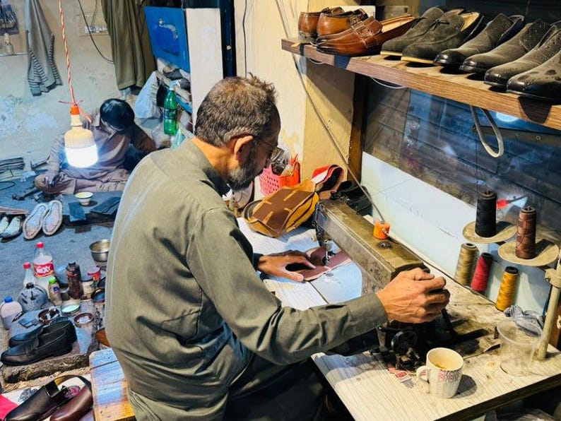 May include: A person in a grey long-sleeved shirt operates a vintage industrial sewing machine to stitch leather pieces. The workspace features various spools of colored thread, finished leather shoes on a shelf, and scattered footwear.