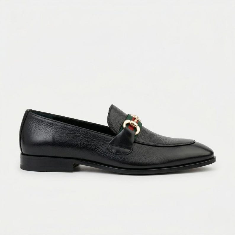 May include: A black leather loafer with a textured finish, featuring a gold-toned metal horsebit detail across the vamp. The shoe includes a green and red fabric stripe accent positioned behind the metal hardware and a low, flat heel.
