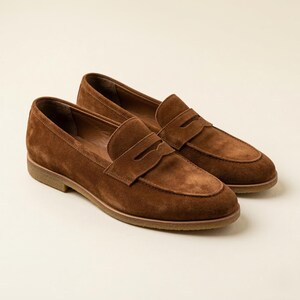 Könnte beinhalten: Ein Paar braune Penny-Loafer aus Wildleder. Die Schuhe haben ein klassisches Design mit runder Spitze, einem Riemen über dem Spann und einem niedrigen Absatz. Die Loafer sind aus weichem, strukturiertem Wildleder.