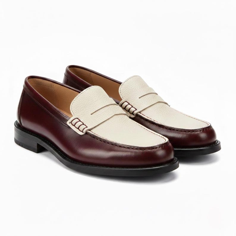 May include: A pair of two-tone loafers featuring a deep burgundy smooth leather heel and side panels paired with a textured cream-colored leather vamp. The shoes include a classic penny loafer strap across the top with visible stitching details.