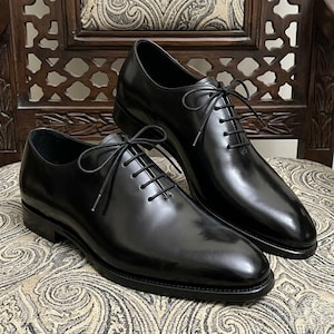Könnte beinhalten: Ein Paar schwarze Oxford-Schuhe aus Leder mit polierter Oberfläche, ausgestattet mit einem geschlossenen Schnürsystem, dünnen schwarzen Schnürsenkeln in Schleifenform, einer abgerundeten Schuhspitze und einem flachen, geschichteten Absatz.