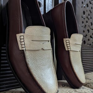 May include: A pair of two-tone loafers featuring dark brown smooth leather sides and a textured cream-colored leather vamp. Each shoe includes a classic penny loafer strap across the top with visible stitching details and a rounded toe design.