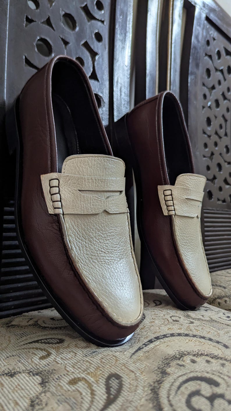 May include: A pair of two-tone loafers featuring dark brown smooth leather sides and a textured cream-colored leather vamp. Each shoe includes a classic penny loafer strap across the top with visible stitching details and a rounded toe design.