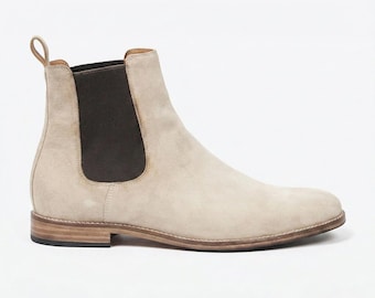 May include: A beige suede Chelsea boot featuring a dark brown elastic side panel, a pull tab at the heel, and a stacked wooden heel with a black rubber sole. The ankle-high boot has a rounded toe and visible stitching along the welt.