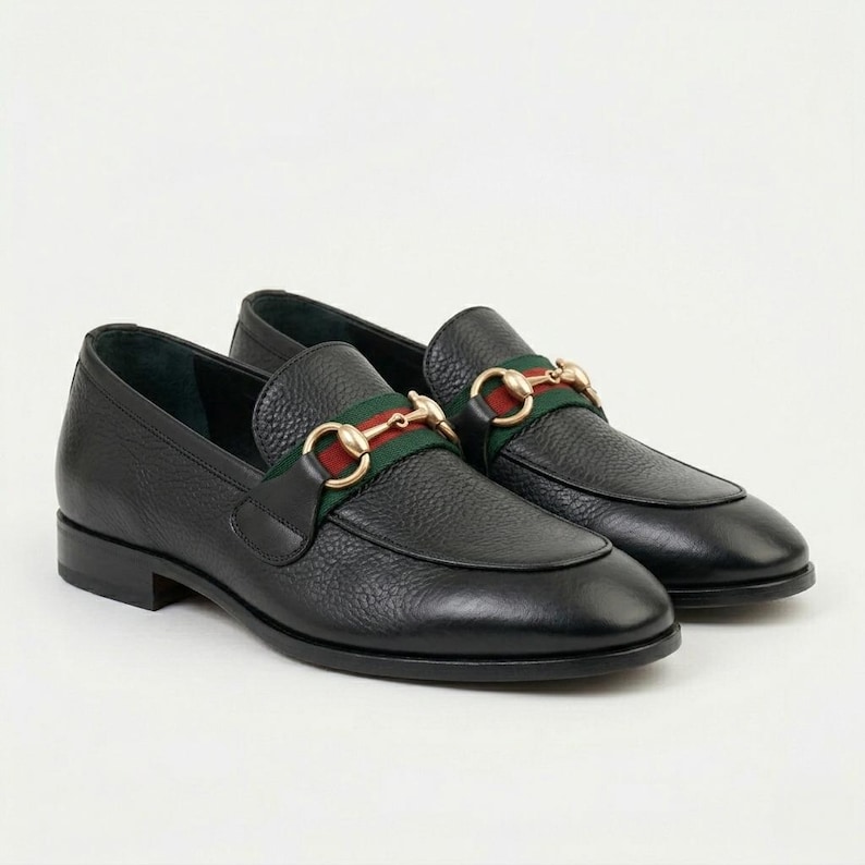 May include: A pair of black leather loafers featuring a textured grain finish. Each shoe is adorned with a green and red fabric web stripe across the vamp, accented by a polished gold-tone metal horsebit hardware detail.
