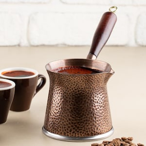 May include: A copper coffee pot with a wooden handle, two brown coffee cups, and a small dish of chocolate squares. Coffee beans are scattered near the pot. The background is a white brick wall.