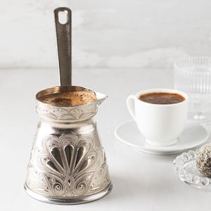 May include: A silver coffee pot with an ornate design, filled with coffee, sits next to a white cup of coffee on a saucer. A glass of water and a small plate with a treat complete the scene, all on a white surface.