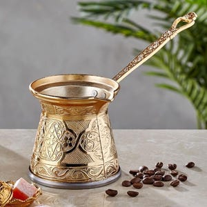 Op de afbeelding: Een goudkleurige, sierlijke koffiepot met een lange handgreep, naast een klein kopje en een schaal met Turks fruit. De koffiepot heeft gedetailleerde bloempatronen. Koffiebonen zijn verspreid over het oppervlak.