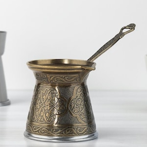 Może przedstawiać: Ozdobny, brązowy turecki dzbanek do kawy, czyli cezve, z długim uchwytem i skomplikowanymi wzorami kwiatowymi. Wewnątrz spoczywa pasująca łyżka. Młynek do kawy ze stali nierdzewnej i biały kubek znajdują się w tle.