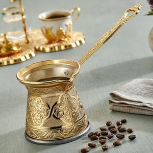 Könnte beinhalten: Eine goldfarbene türkische Kaffeekanne mit aufwendigen floralen Mustern, einem langen Griff und einem kleinen Ausguss. Die Kanne steht neben einem passenden Tassen- und Untertassenset, einem kleinen Glashalter und einem Haufen Kaffeebohnen. Ein gefaltetes Leinentuch und eine Vase mit Blumen sind ebenfalls im Bild.