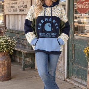 May include: A color-blocked hooded sweatshirt featuring a floral print on the shoulders and sleeves, a navy blue chest panel with the text Carhartt Made For Work Established 1889, olive green side panels, and a light blue kangaroo pocket.