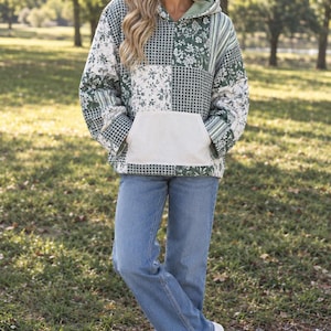 May include: A long-sleeve hooded sweatshirt featuring a patchwork design of green and white floral, striped, and checkered patterns. The garment includes a solid cream-colored front kangaroo pocket, paired with light blue denim jeans and white sneakers.