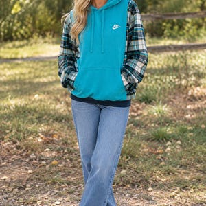 May include: A teal hooded sweatshirt featuring a white Nike logo on the chest, long sleeves with a green, white, and black plaid pattern, a front kangaroo pocket, and a dark blue hem, paired with light blue denim jeans and white sneakers.