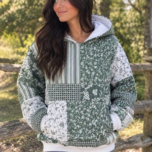 May include: A quilted hoodie featuring a patchwork design of green and white floral, striped, and checkered patterns. The garment includes a white hood lining, white ribbed cuffs, a white bottom hem, and a large front kangaroo pocket.