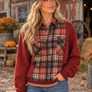 May include: A dark red long-sleeve sweatshirt featuring a central plaid button-up shirt panel with red, black, and white patterns, two chest pockets, and a ribbed hem with white stripes, paired with a distressed black baseball cap.