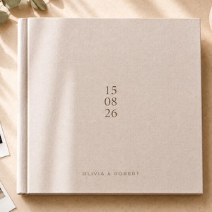 May include: A square, light beige fabric-covered photo album featuring the date 15 08 26 printed in the center and the names OLIVIA & ROBERT printed at the bottom in a dark, minimalist sans-serif font on the front cover.