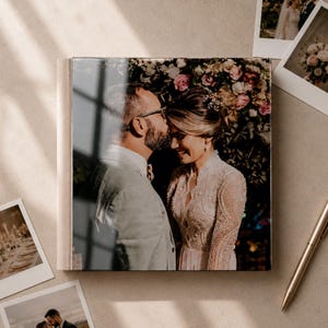 Puede incluir: Un álbum de fotos cuadrado con una cubierta brillante que muestra a una pareja con ropa formal, donde la persona de la izquierda besa la frente de la persona de la derecha. El álbum está rodeado de fotos estilo polaroid y un bolígrafo dorado.