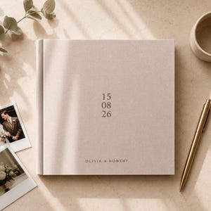 Puede incluir: Un álbum de fotos cuadrado cubierto de tela beige claro con la fecha 15 08 26 y los nombres OLIVIA & ROBERT impresos en una fuente oscura y minimalista en la portada, acompañado de un bolígrafo dorado y dos fotos cuadradas pequeñas.