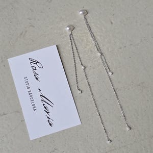 Puede incluir: Un par de pendientes colgantes de tono plateado con pequeñas perlas blancas en la parte superior y cadenas delicadas con diminutos detalles de perlas. Incluye una tarjeta rectangular blanca con el texto impreso Rosa Maria STUDIO BARCELONA.