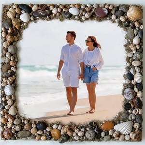 Op de afbeelding: Een vierkante fotolijst met een rand van schelpen en kiezels. De lijst omringt een foto van een stel dat op het strand loopt. De natuurlijke elementen van de lijst creëren een kustthema.