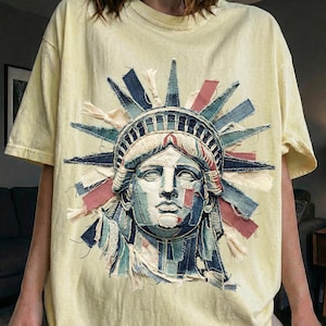 Op de afbeelding: Lichtgele t-shirt met een afbeelding van het Vrijheidsbeeld. De afbeelding is gemaakt van stoffen patches in blauw, rood en crème. Het shirt heeft korte mouwen en een ronde hals.