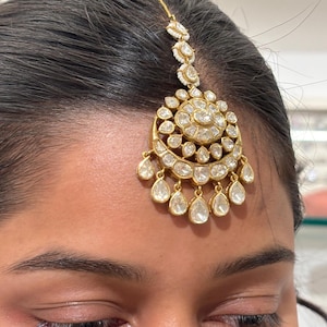 May include: A gold-toned headpiece with a central circular design and dangling teardrop-shaped accents. The headpiece is adorned with numerous clear, sparkling stones, creating a radiant effect. The piece is designed to be worn on the forehead.