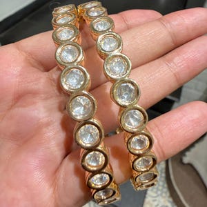 Può includere: Due bracciali rigidi color oro caratterizzati da una fila continua di pietre circolari, trasparenti e sfaccettate, incastonate in singole montature metalliche. I bracciali presentano una finitura metallica lucida e un design uniforme.