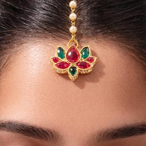 May include: A gold-toned headpiece with a lotus flower design, adorned with red, green, and clear gemstones. The floral centerpiece is suspended from a strand of pearls, creating an elegant and eye-catching accessory.