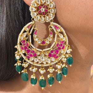 May include: Ornate gold-coloured earrings with a floral and crescent moon design. The earrings feature clear and pink gemstones, white pearls, and dangling green beads. The earrings are decorative and striking.