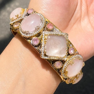 Può includere: Un bracciale rigido color oro con grandi pietre cabochon lisce color rosa pallido, di forma ovale e quadrata. Il bracciale è arricchito da piccole pietre rosa rotonde e incastonato con minuscoli cristalli trasparenti lungo le montature.