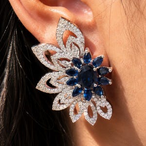 May include: A large statement earring featuring a central oval-cut dark blue gemstone surrounded by a halo of pear-cut blue stones. The design includes an asymmetrical fan-shaped section covered in small, clear pavé-set white crystals.