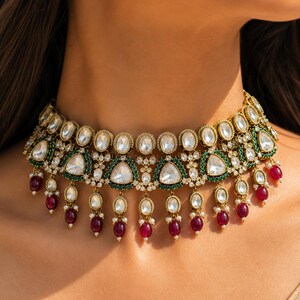 May include: A gold-toned choker necklace featuring a central row of oval white stones, triangular white stones framed by small green beads, and dangling teardrop-shaped red stones accented with tiny white pearls along the bottom edge.