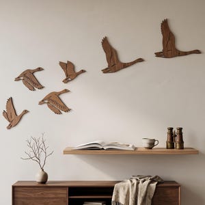 May include: A set of five brown wooden wall art pieces shaped like flying geese, arranged in a flock formation. Each bird silhouette features cut-out wing details, showcasing a natural wood grain texture and a smooth, flat finish on the surface.