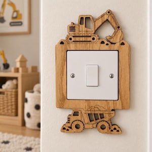 May include: A light switch cover plate made of light-coloured wood featuring a laser-cut design of an excavator at the top and a dump truck at the bottom. The central square plate is white with a single vertical toggle switch and two silver screws.