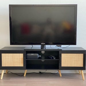 May include: A black TV stand with two doors featuring woven cane panels and light wood legs. A black Samsung television sits on top. A black fan and a potted snake plant are visible.