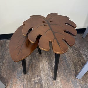 May include: Two brown leaf-shaped side tables. The tables have black legs and are made of wood. The larger table is in the shape of a monstera leaf. The smaller table is oval-shaped. The tables are a decorative accent for a living room or bedroom.