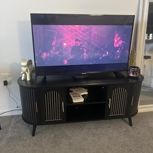 Pode incluir: M&oacute;vel de TV preto com detalhes dourados, suportando uma grande televis&atilde;o de tela plana. O arm&aacute;rio tem duas portas e uma prateleira, com o texto "THINK RICH" em um livro. Uma est&aacute;tua decorativa de elefante e um pote de vidro est&atilde;o em cima.