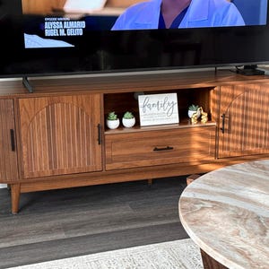 Pode incluir: Um m&oacute;vel de TV em estilo moderno de meados do s&eacute;culo, com acabamento castanho quente. O m&oacute;vel possui duas portas de arm&aacute;rio com detalhes de ranhuras verticais, uma gaveta e uma prateleira aberta. Uma placa com a palavra "family" est&aacute; na prateleira, com pequenas plantas em vasos.
