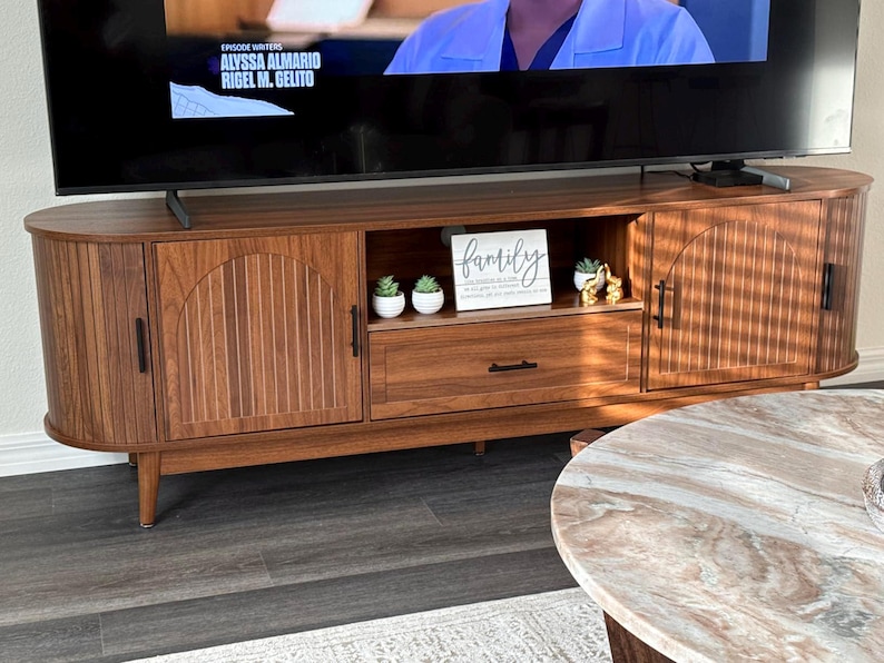 Pode incluir: Um m&oacute;vel de TV em estilo moderno de meados do s&eacute;culo, com acabamento castanho quente. O m&oacute;vel possui duas portas de arm&aacute;rio com detalhes de ranhuras verticais, uma gaveta e uma prateleira aberta. Uma placa com a palavra "family" est&aacute; na prateleira, com pequenas plantas em vasos.