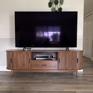 Pode incluir: Um m&oacute;vel de TV de madeira em estilo moderno de meados do s&eacute;culo com uma grande televis&atilde;o de tela preta em cima. O m&oacute;vel tem duas portas, uma gaveta e uma prateleira aberta. Um console de videogame e um controle s&atilde;o vis&iacute;veis.