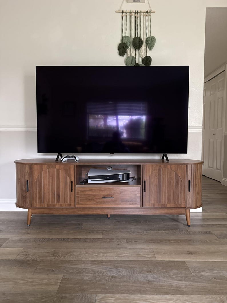 Pode incluir: Um m&oacute;vel de TV de madeira em estilo moderno de meados do s&eacute;culo com uma grande televis&atilde;o de tela preta em cima. O m&oacute;vel tem duas portas, uma gaveta e uma prateleira aberta. Um console de videogame e um controle s&atilde;o vis&iacute;veis.