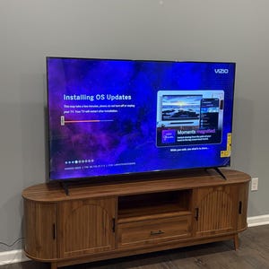 Pode incluir: Uma TV Vizio exibindo "Installing OS Updates" em uma consola de m&iacute;dia de madeira. A tela mostra um fundo azul e roxo com o logotipo da Vizio. A consola tem duas portas, uma gaveta e um design curvo.