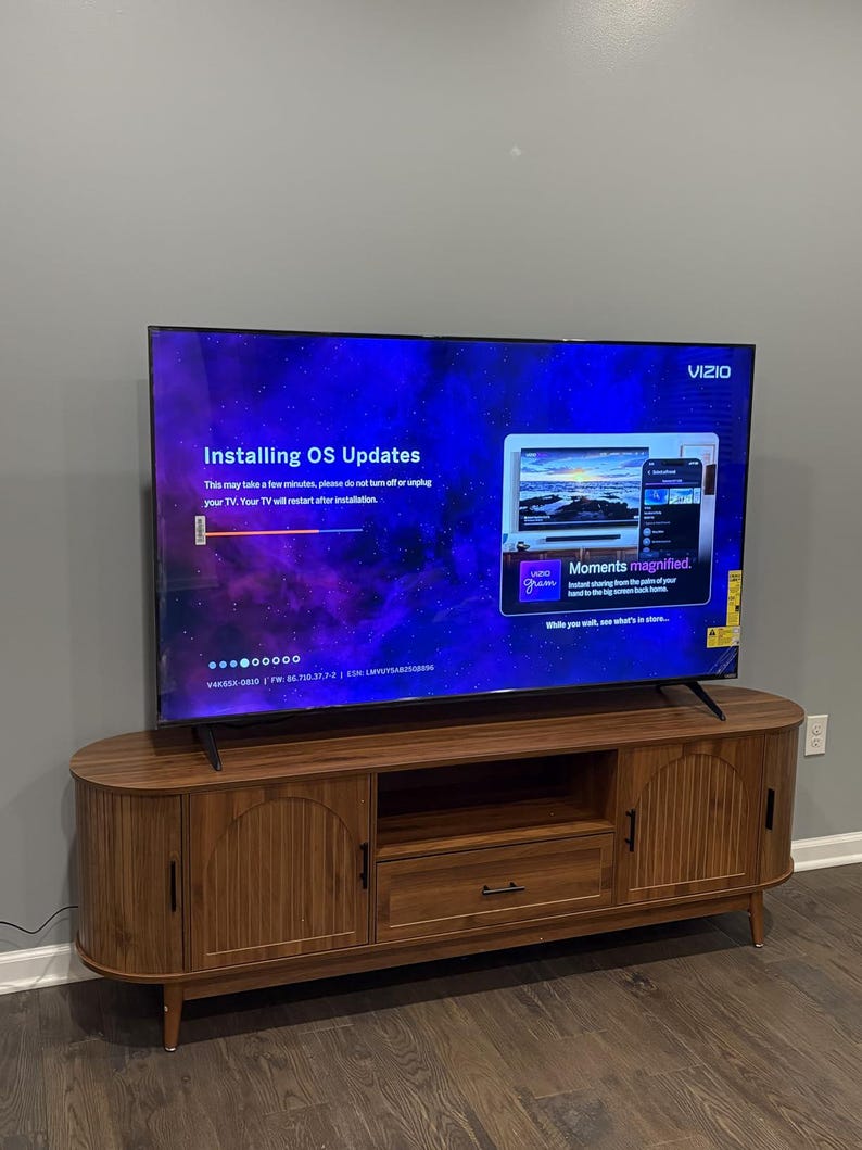 Pode incluir: Uma TV Vizio exibindo "Installing OS Updates" em uma consola de m&iacute;dia de madeira. A tela mostra um fundo azul e roxo com o logotipo da Vizio. A consola tem duas portas, uma gaveta e um design curvo.