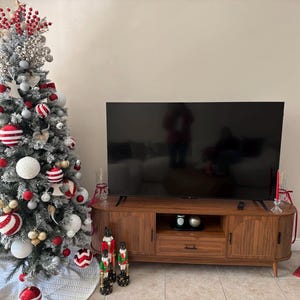 Pode incluir: Uma &aacute;rvore de Natal decorada com enfeites vermelhos, brancos e prateados est&aacute; ao lado de um m&oacute;vel de TV de madeira. A &aacute;rvore &eacute; flocada de branco e tem uma variedade de enfeites. Uma grande TV de tela plana est&aacute; montada acima.