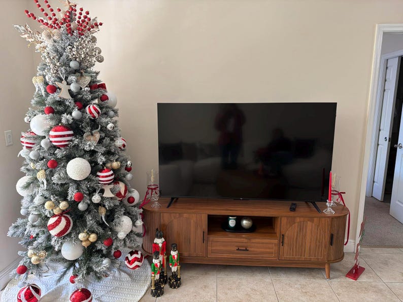 Pode incluir: Uma &aacute;rvore de Natal decorada com enfeites vermelhos, brancos e prateados est&aacute; ao lado de um m&oacute;vel de TV de madeira. A &aacute;rvore &eacute; flocada de branco e tem uma variedade de enfeites. Uma grande TV de tela plana est&aacute; montada acima.