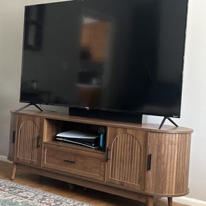 Pode incluir: Um m&oacute;vel de TV de madeira com um design moderno de meados do s&eacute;culo. O m&oacute;vel tem duas portas com ranhuras verticais, uma gaveta e uma prateleira. Uma grande televis&atilde;o preta est&aacute; em cima. O m&oacute;vel &eacute; castanho com ferragens pretas.
